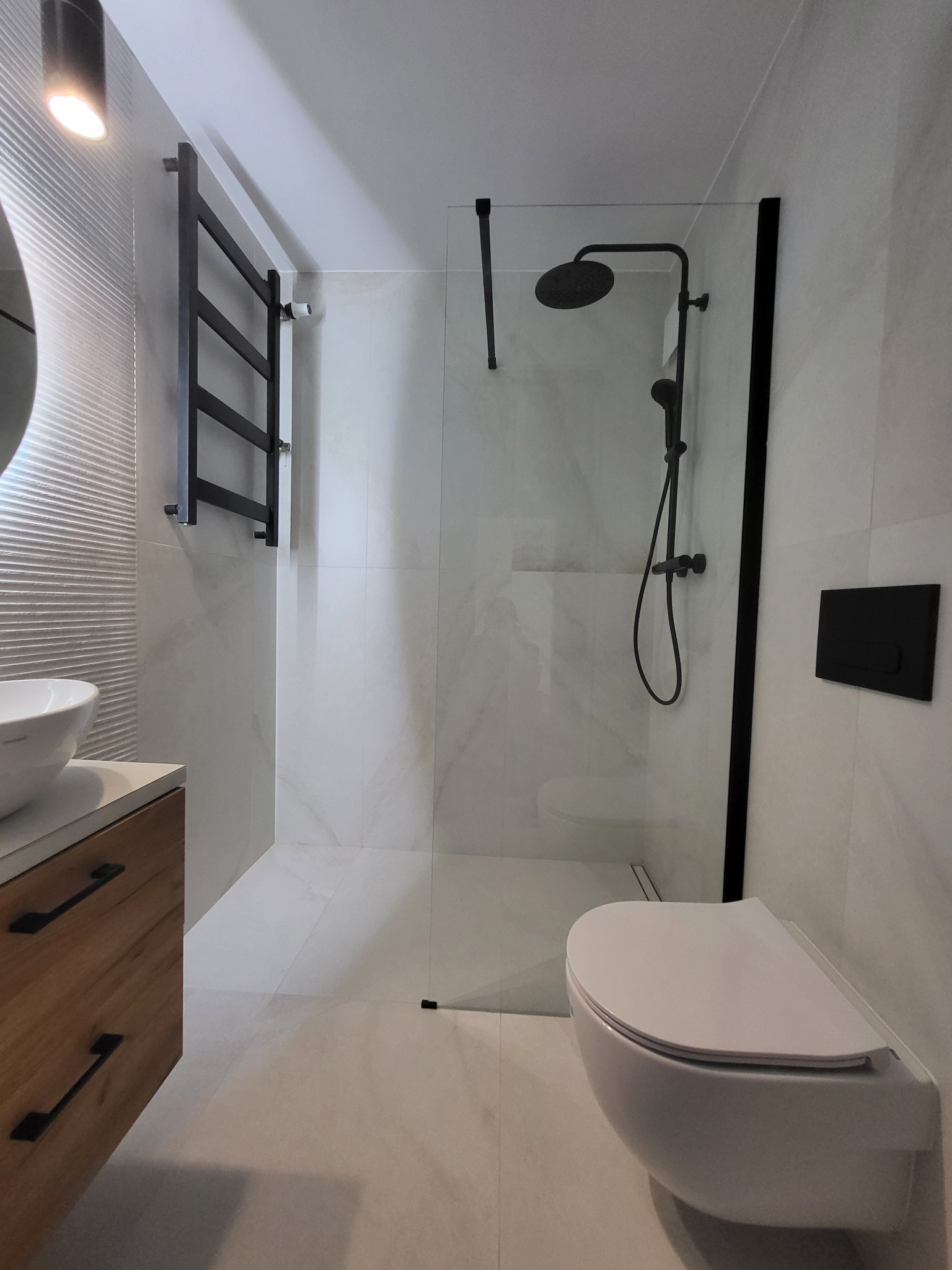 Minimalistyczna łazienka z kabiną typu walk-in, czarną armaturą i drewnianą szafką pod umywalką. Ściany i podłoga wyłożone płytkami imitującymi marmur.