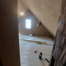 Poddasze w trakcie prac wykończeniowych: świeżo otynkowane ściany, przygotowane deski podłogowe, narzędzia i akcesoria rozłożone na podłodze, widoczny fragment drewnianej belki konstrukcyjnej.