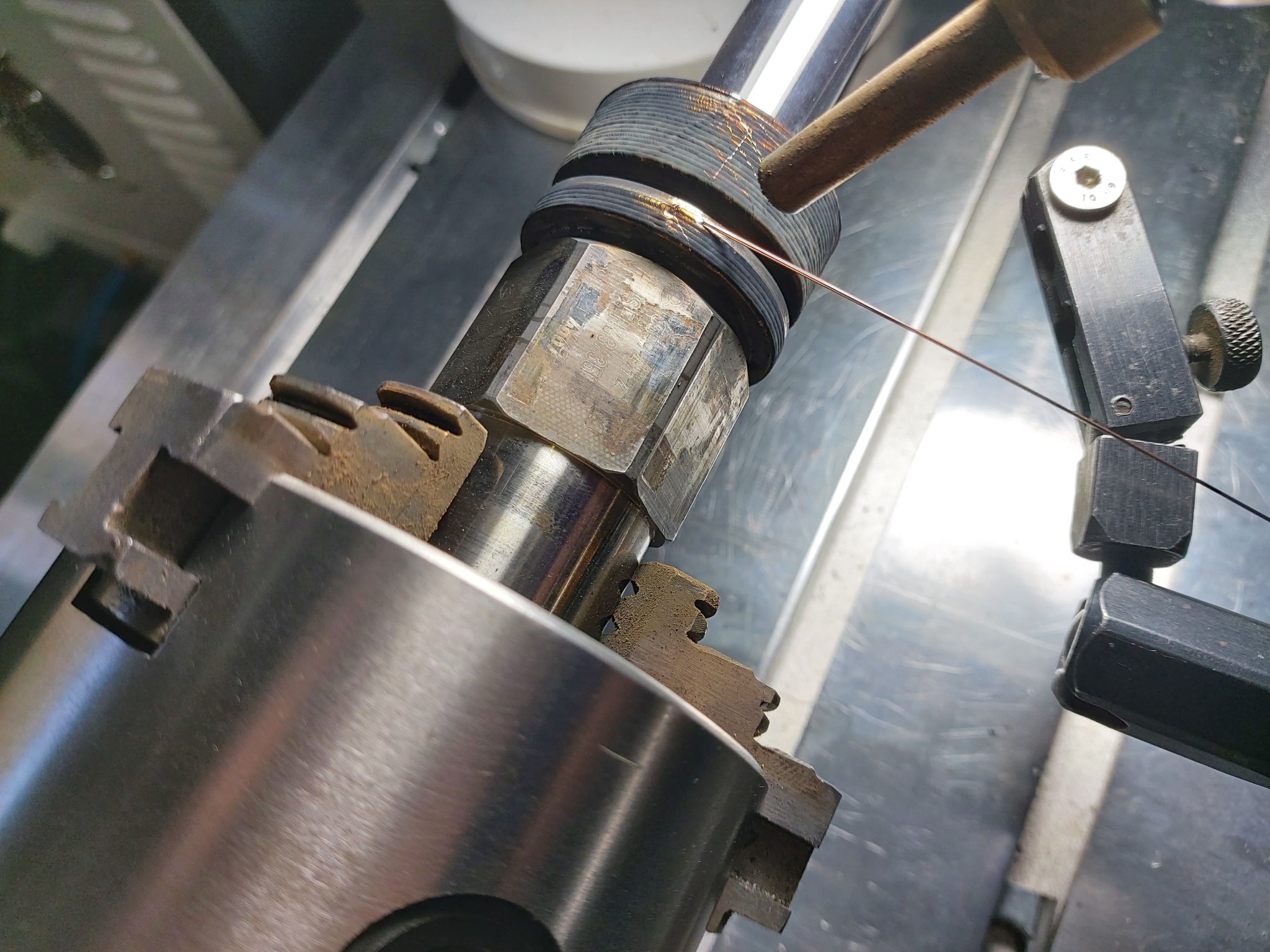 Precyzyjne spawanie laserowe cienkiego drutu do metalowego elementu mocowanego w uchwycie tokarskim, widoczne delikatne iskry i ślady obróbki na powierzchni metalu.