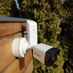 Zewnętrzna kamera monitoringu Hikvision zamontowana na drewnianej ścianie budynku, obok widoczny moduł komunikacyjny Unifi, tło stanowią zielone krzewy.