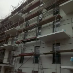 Wielopiętrowy budynek w trakcie prac wykończeniowych, osłonięty rusztowaniem, widoczne okna zabezpieczone folią i pojemniki z farbą na balkonach.