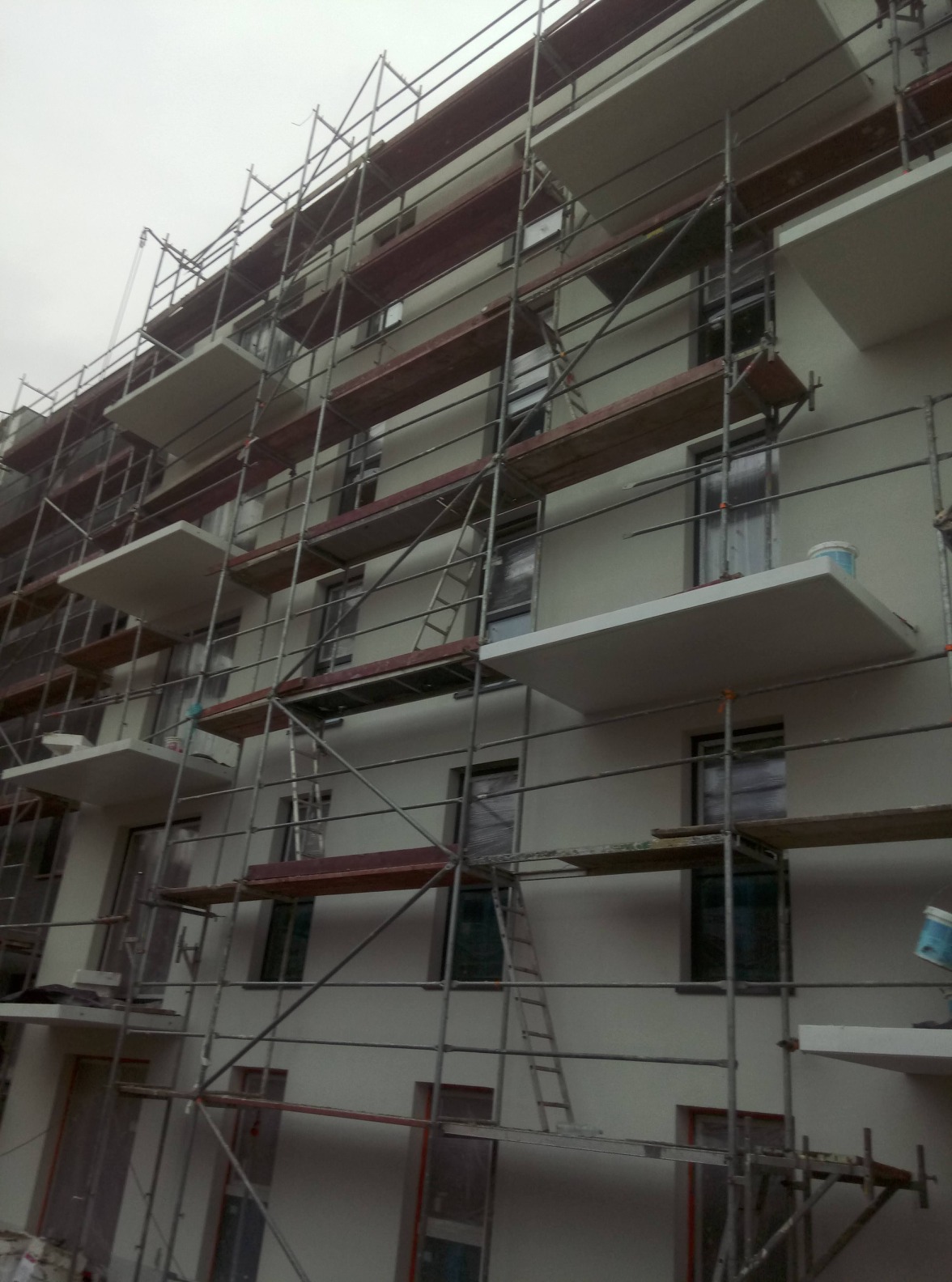 Wielopiętrowy budynek w trakcie prac wykończeniowych, osłonięty rusztowaniem, widoczne okna zabezpieczone folią i pojemniki z farbą na balkonach.
