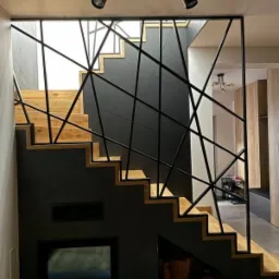 Schody drewniane z czarną, geometryczną balustradą i czarną ścianą z wnęką, oświetlone reflektorami na szynie.