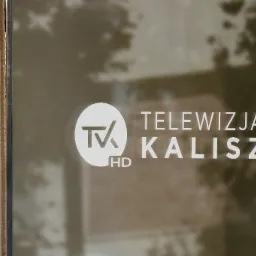 Białe logo 'Telewizja Kalisz HD' na szklanej powierzchni z odbiciem otoczenia, przylegające do kolumny z piaskowca.