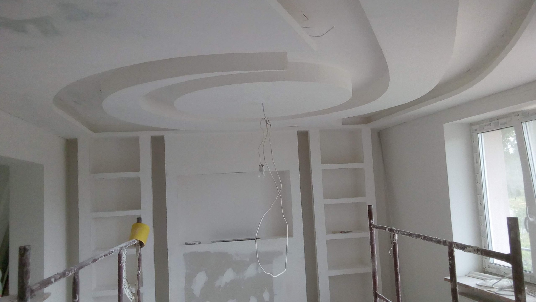 Białe, okrągłe, gipsowo-kartonowe wykończenie sufitu z wbudowanymi półkami w ścianie, widoczne z rusztowania w trakcie prac wykończeniowych.