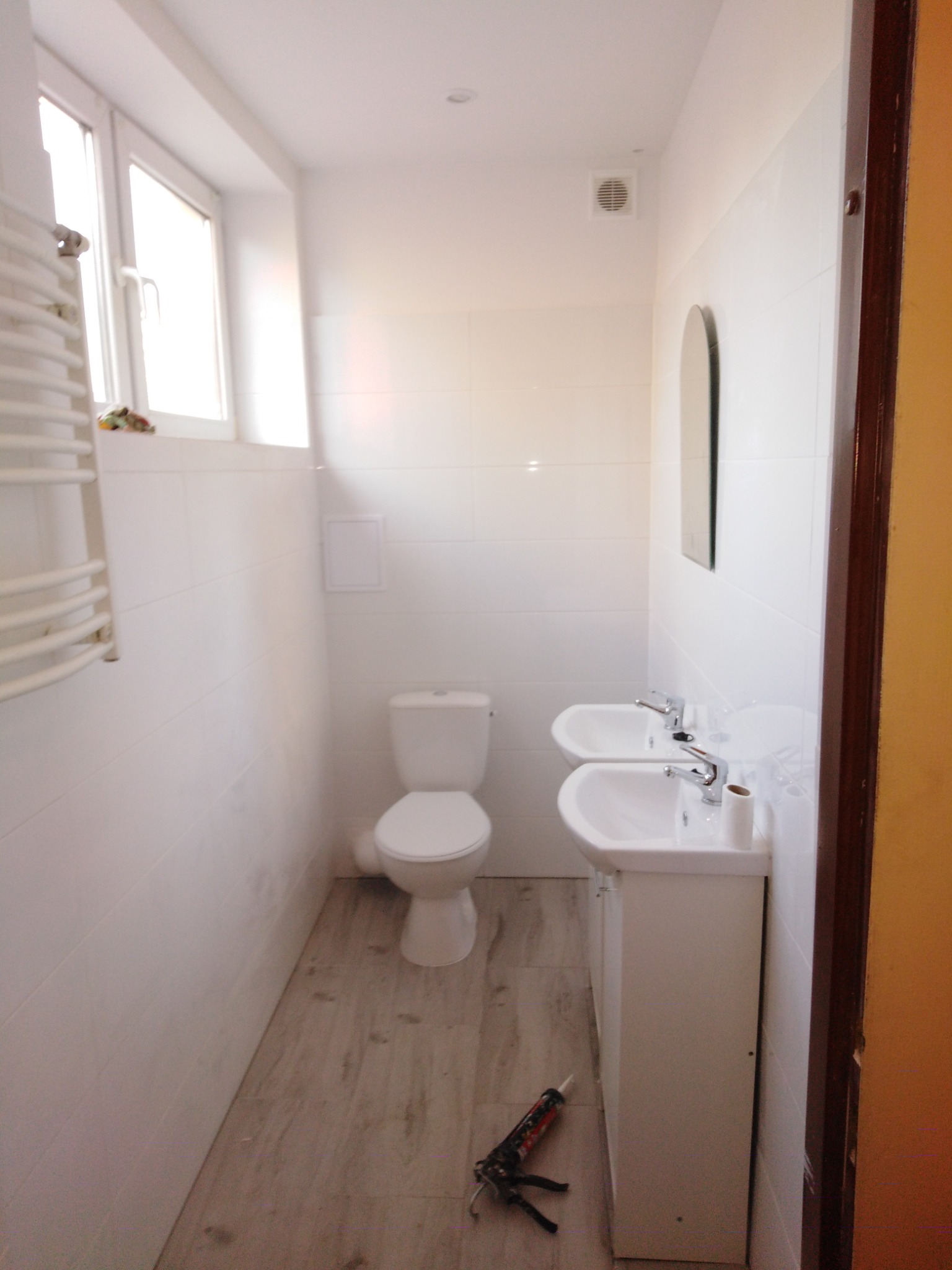 Wnętrze łazienki w trakcie prac: białe płytki, dwie umywalki z bateriami, toaleta, grzejnik drabinkowy, drewnopodobna podłoga, pistolet do silikonu na podłodze.