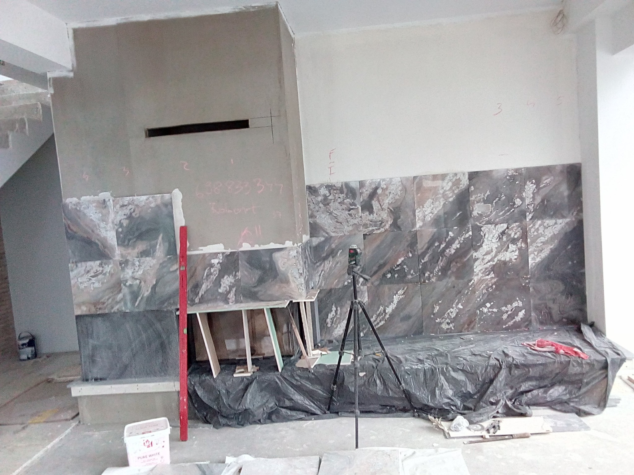 Montaż płytek imitujących marmur na ścianie kominka w trakcie prac, widoczny laser krzyżowy na statywie, poziomica, wiadro z farbą i notatki na ścianie.