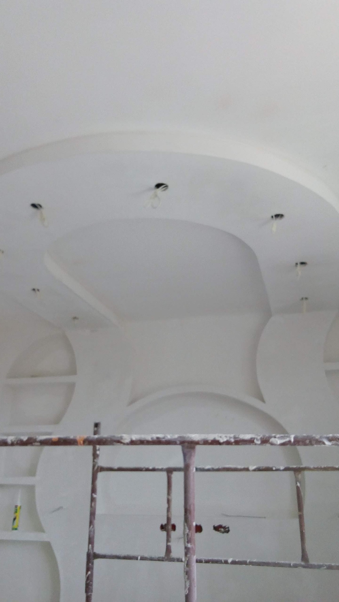 Biały sufit z dekoracyjnymi elementami gipsowymi w kształcie łuków i okręgów, widoczne otwory na oświetlenie, rusztowanie malarskie w dolnej części kadru.