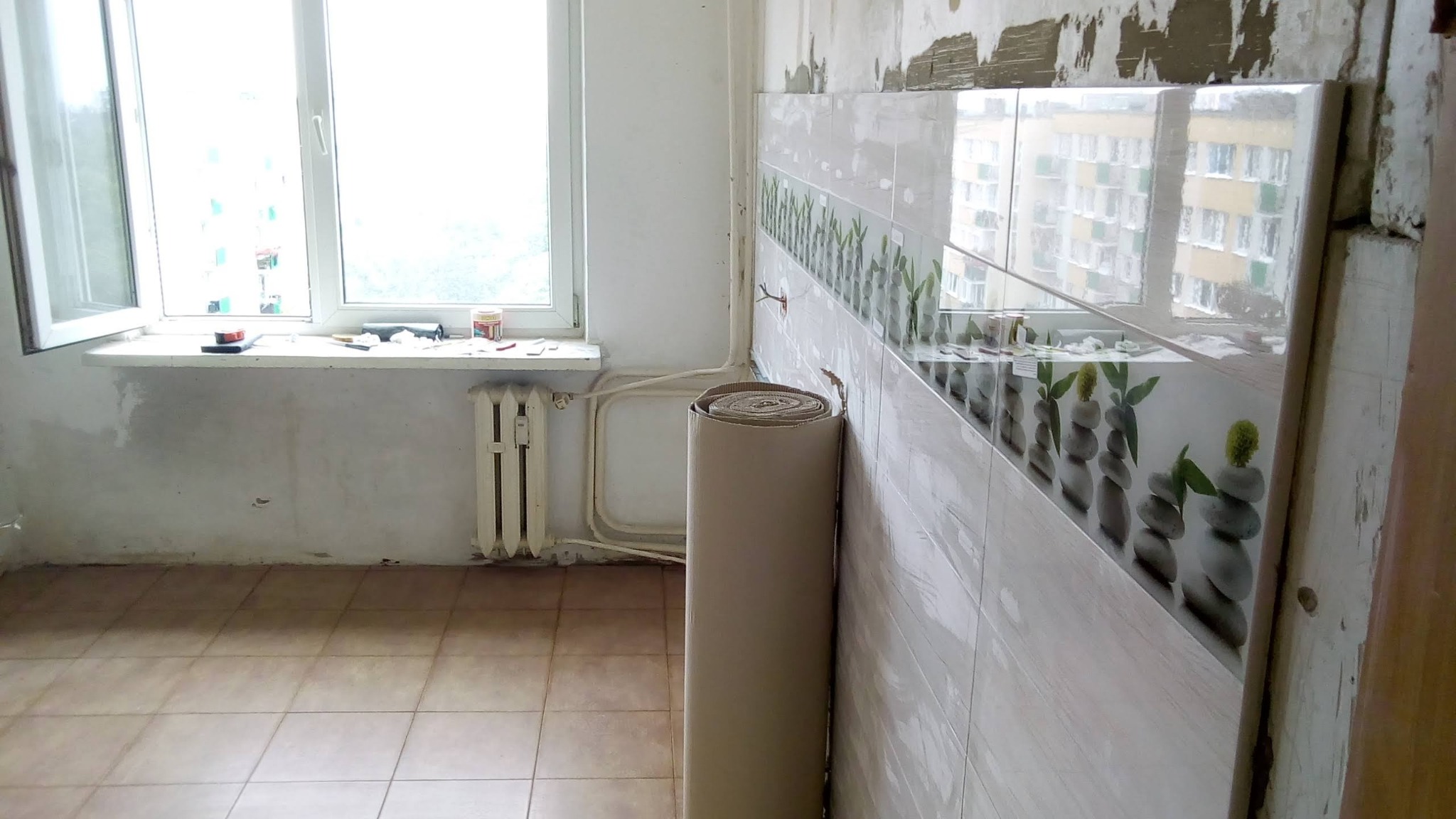 Przygotowania do prac w kuchni: rolka wykładziny podłogowej oparta o ścianę z częściowo położonymi płytkami z motywem zen, widoczne okno i grzejnik w tle.