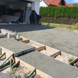 Świeżo wylana betonowa posadzka z drewnianym szalunkiem na zewnątrz, widok na garaż i ogrodzenie z adresem.