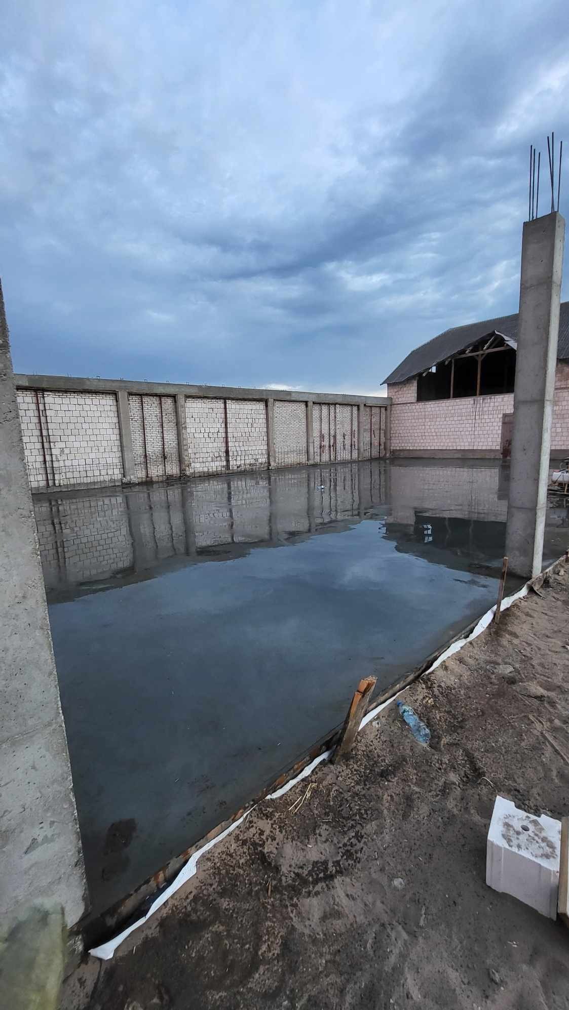 Świeżo wylana, mokra posadzka na budowie, z widocznymi elementami konstrukcyjnymi budynku w tle i pochmurnym niebem.