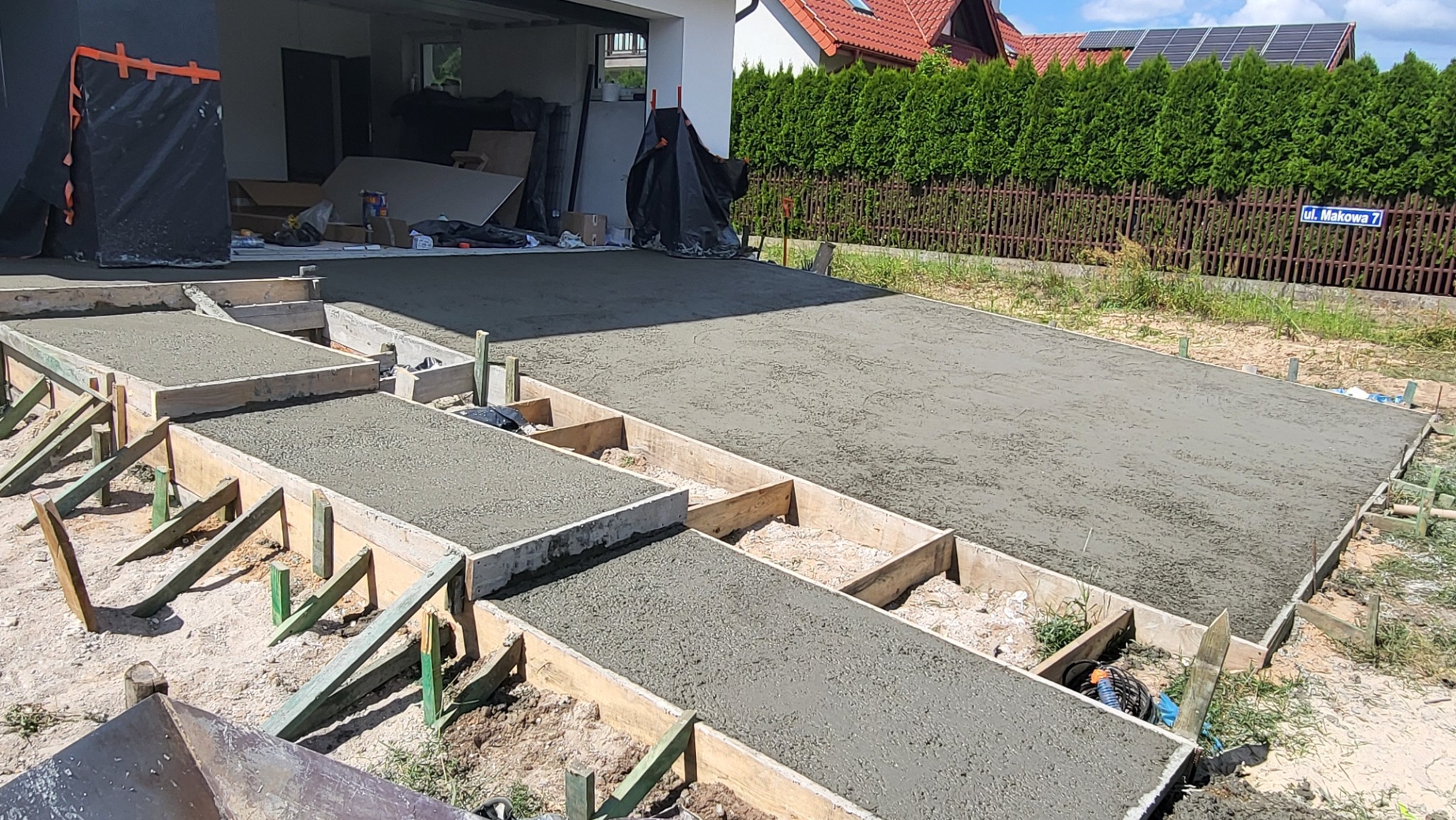 Świeżo wylana betonowa posadzka z drewnianym szalunkiem na zewnątrz, widok na garaż i ogrodzenie z adresem.