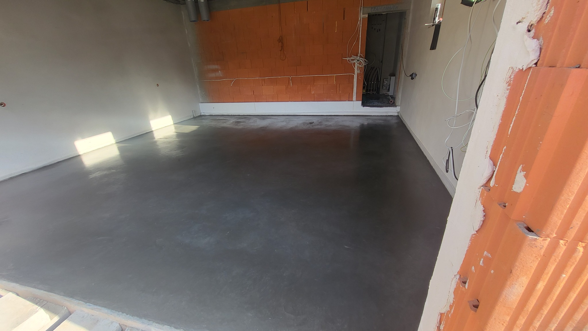 Świeżo wylana, gładka posadzka w surowym wnętrzu z widocznymi instalacjami i niedokończonymi ścianami z cegły.