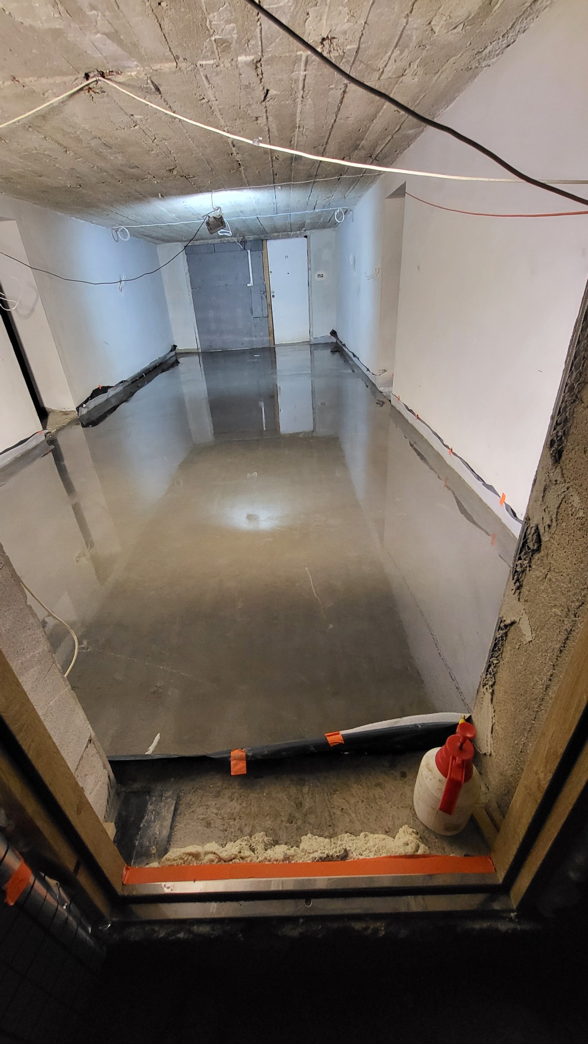 Świeżo wylana, błyszcząca posadzka w pomieszczeniu z surowym betonowym sufitem i białymi ścianami, widoczne zabezpieczenia przyścienne i plastikowa butelka z czerwoną pompką w rogu.