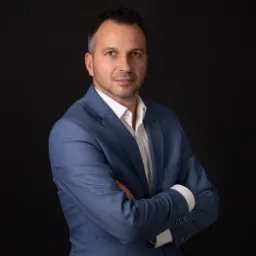 Portret mężczyzny w błękitnym garniturze i białej koszuli, skrzyżowane ramiona na tle czarnego tła.