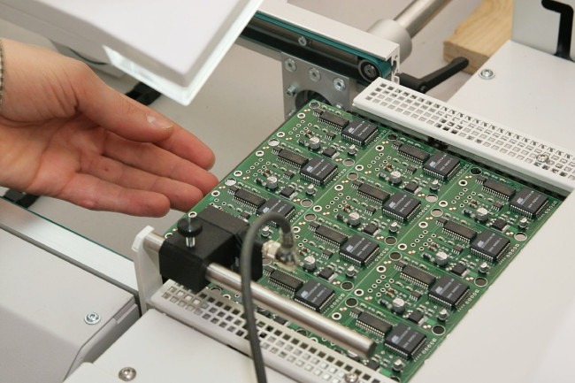 Zielona płytka drukowana z komponentami elektronicznymi umieszczona w automatycznym urządzeniu, dłoń osoby w lewym górnym rogu, prawdopodobnie podczas procesu produkcyjnego lub testowania.