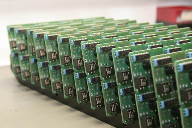 Rząd zielonych płytek drukowanych z czarnymi elementami elektronicznymi, ustawionych pionowo w rzędzie na czarnym podłożu, widok z boku pod kątem.