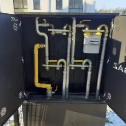 Zewnętrzna szafka gazowa z otwartymi drzwiczkami, widoczne rury gazowe, zawory z żółtymi uchwytami oraz licznik gazu na tle nowoczesnego budynku.