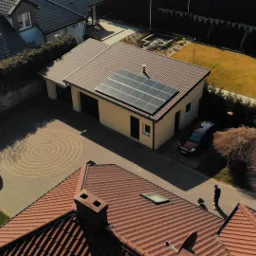 Widok z góry na dom z panelami słonecznymi na dachu garażu, obok zaparkowany samochód, na pierwszym planie fragment dachu innego domu i postać ludzka na trawniku.