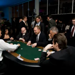 Grupa elegancko ubranych osób gra w pokera przy stole kasynowym podczas imprezy firmowej, w tle choinka i okna.