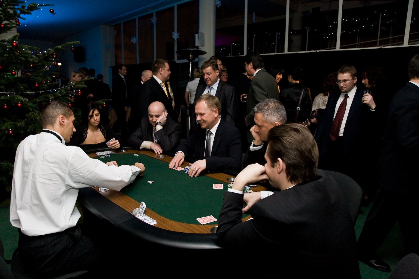 Grupa elegancko ubranych osób gra w pokera przy stole kasynowym podczas imprezy firmowej, w tle choinka i okna.