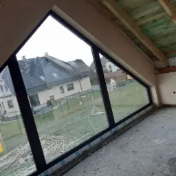 Wykończone tynki przy trójkątnym oknie na poddaszu, widoczne drewniane elementy konstrukcji dachu i widok na dom z ogrodem za oknem.