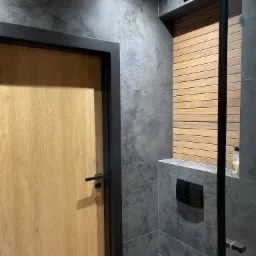 Nowoczesna łazienka z drewnianymi akcentami: jasne drzwi z czarną ościeżnicą, szare płytki imitujące kamień, drewniana boazeria w strefie prysznica, czarna toaleta podtynkowa.