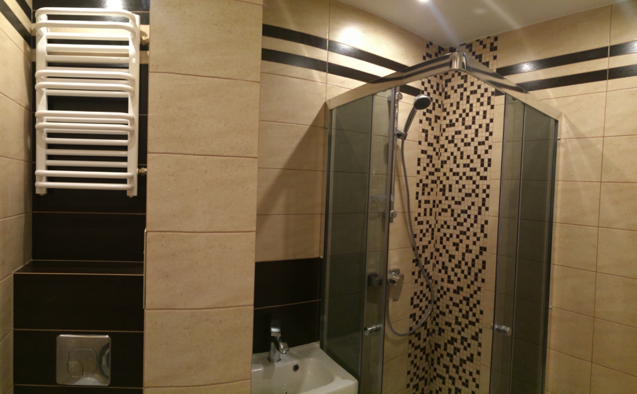 Wykończona łazienka z kabiną prysznicową narożną, grzejnikiem drabinkowym i umywalką w neutralnej kolorystyce płytek z ciemnymi akcentami.