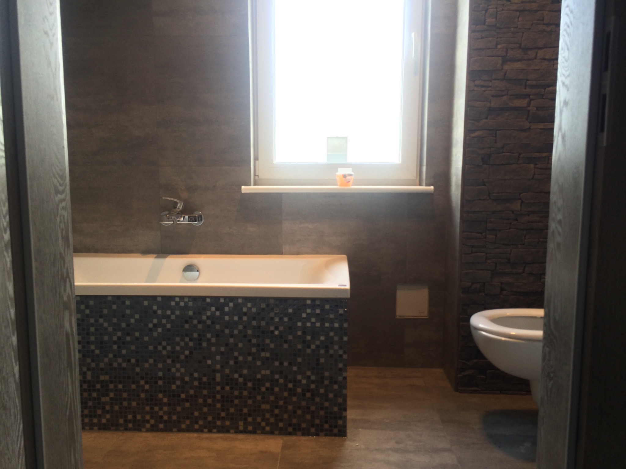 Nowoczesna łazienka z ciemnymi płytkami imitującymi kamień na jednej ścianie, mozaiką na obudowie wanny, i wiszącą toaletą, widziana zza uchylonych drzwi.