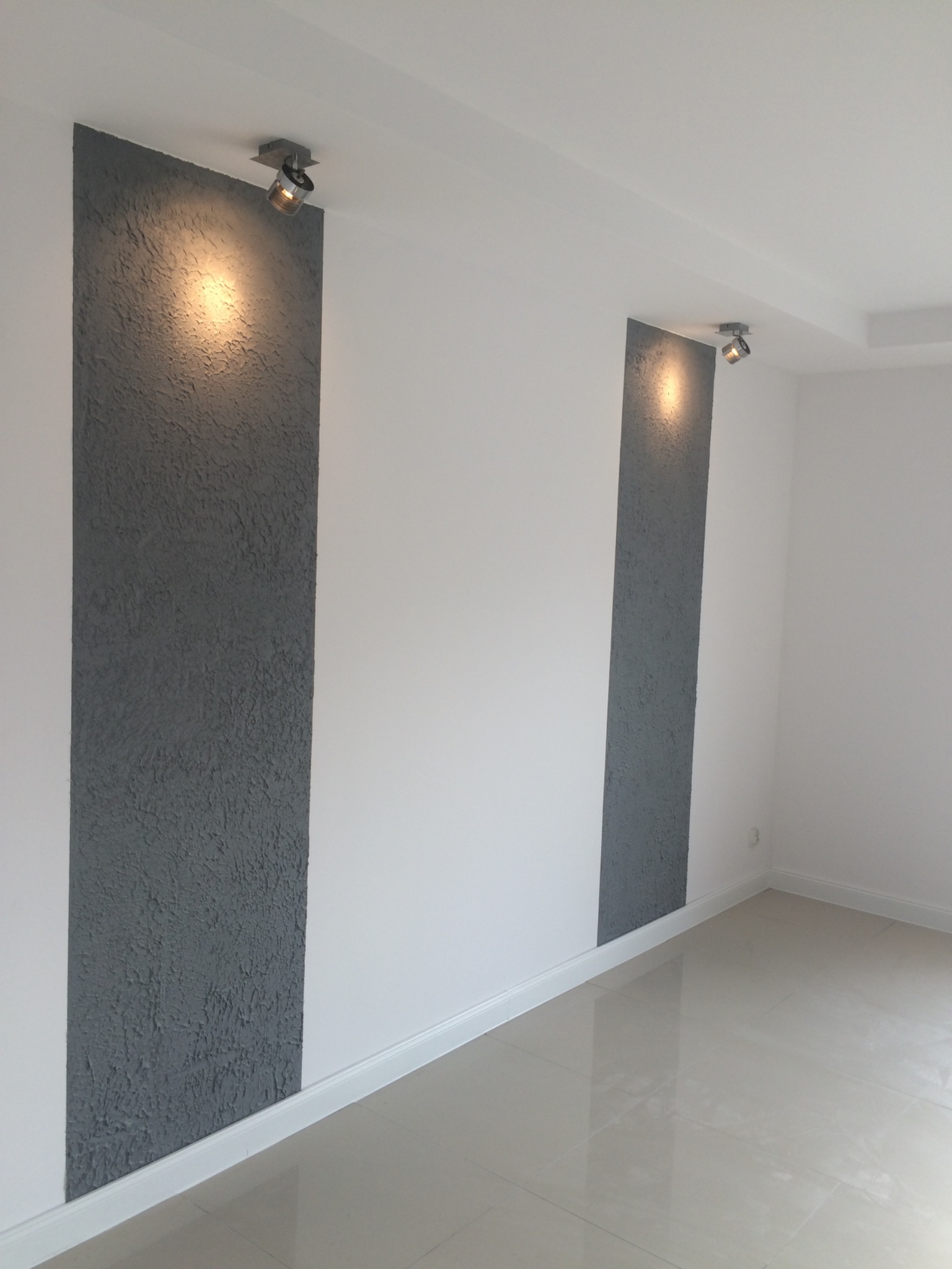 Wnętrze pokoju z wykończonymi ścianami: pionowe pasy szarej, teksturowanej ściany naprzemiennie z białymi, gładkimi powierzchniami. Oświetlenie punktowe podkreśla strukturę. Podłoga wyłożona...