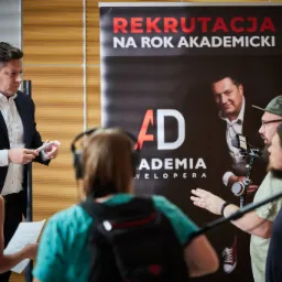 Ekipa filmowa kręci ujęcie z mężczyzną w garniturze, w tle plakat z napisem 'Rekrutacja na rok akademicki' i wizerunkiem mężczyzny.