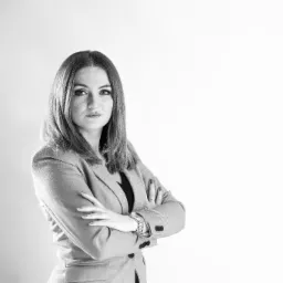 Portret kobiety w szarym garniturze, z założonymi rękami, na jasnym tle, w czerni i bieli, z widocznym zegarkiem na ręce.