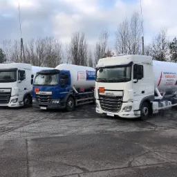 Trzy ciężarówki cysterny z logo Gazgrod zaparkowane na betonowym placu, transportujące propan LPG, widok z przodu na tle drzew bez liści i pochmurnego nieba.