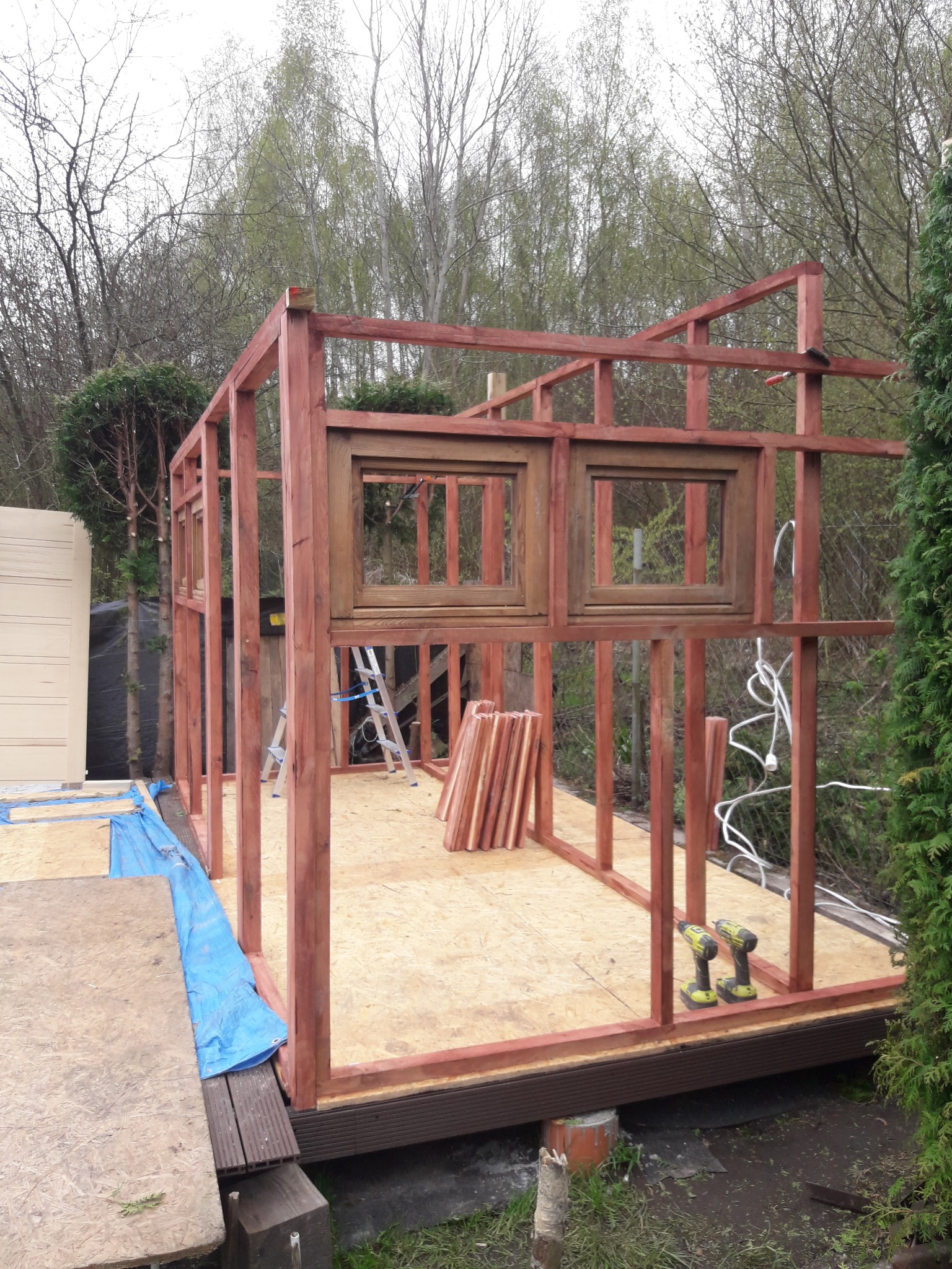 Konstrukcja szkieletowa z drewna, prawdopodobnie altany lub domku letniskowego, z wstawionymi oknami, widoczne narzędzia, drabina i przygotowane elementy drewniane.