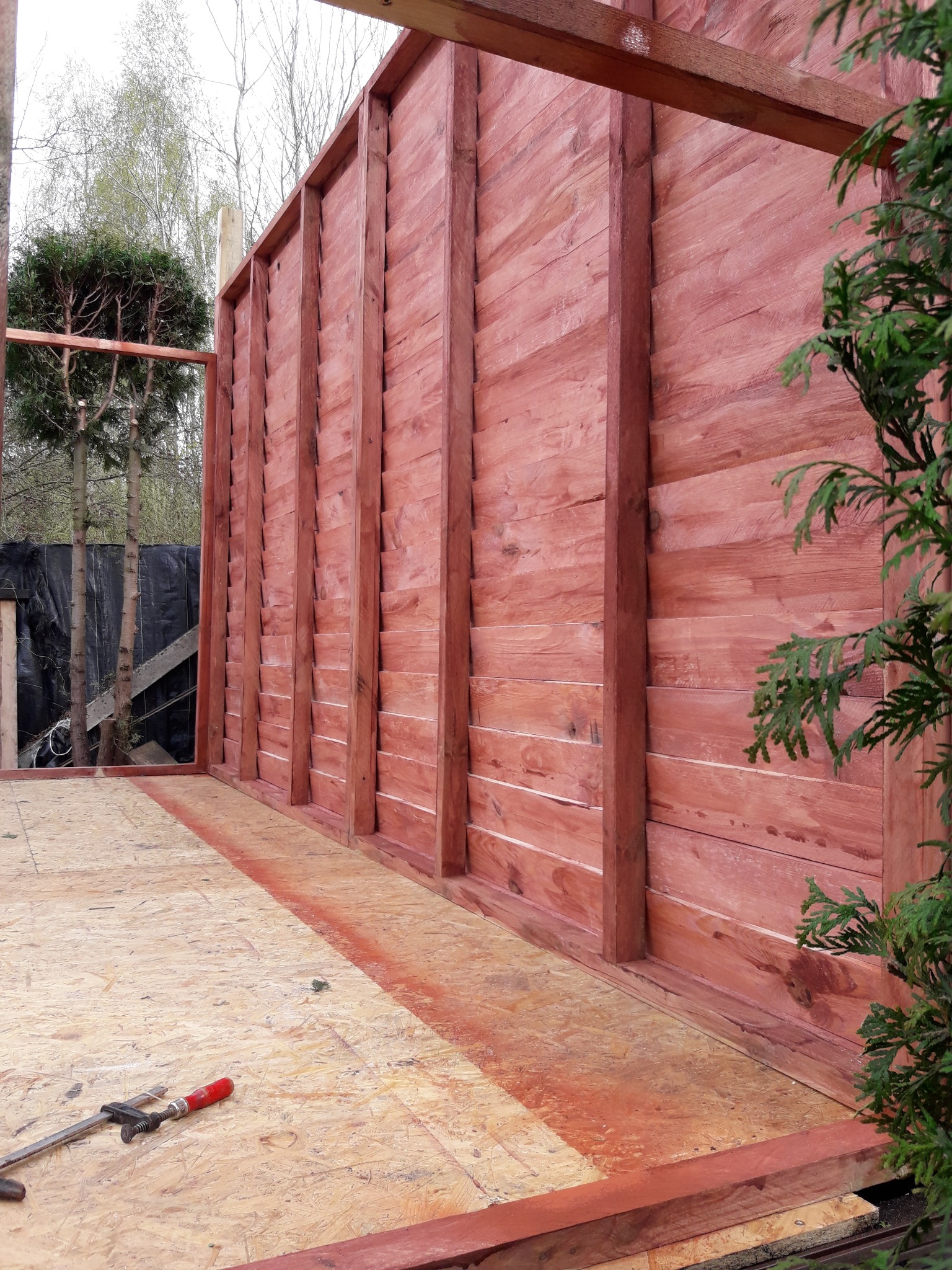 Wnętrze w trakcie budowy drewnianej konstrukcji, prawdopodobnie altany lub domku, z widocznymi deskami pomalowanymi na czerwono-brązowy kolor, podłogą z płyt OSB i narzędziem stolarskim leżącym...