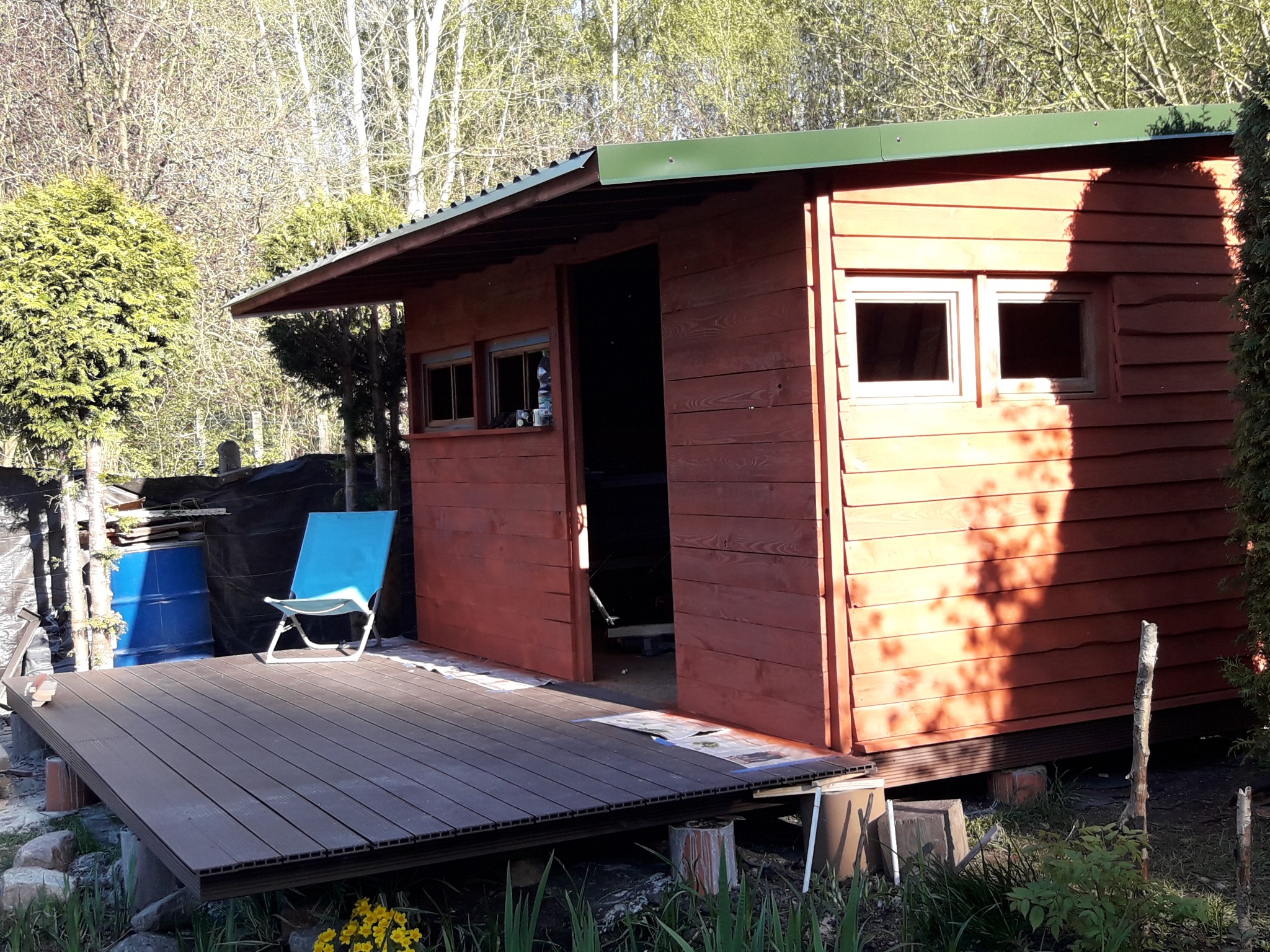 Drewniana altana ogrodowa z zielonym dachem i tarasem z desek kompozytowych, błękitne krzesło turystyczne na tarasie, w tle drzewa i krzewy.