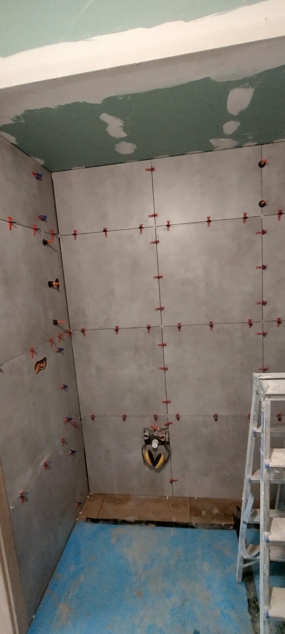 Szare płytki ścienne w trakcie układania w łazience, widoczne krzyżyki dystansowe i poziomica w gniazdku instalacyjnym, niebieska folia w płynie na podłodze, drabina aluminiowa w rogu.