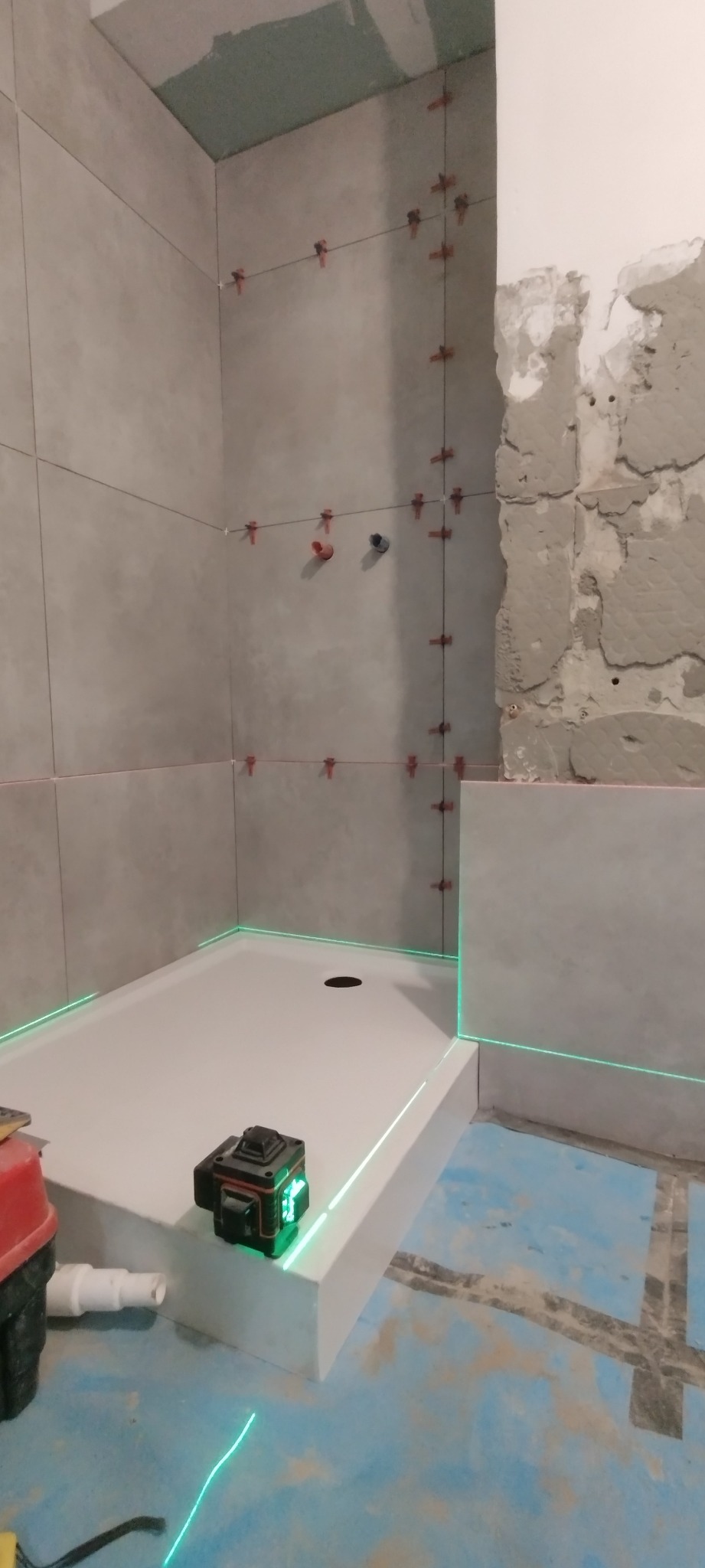 Montaż płytek w łazience z użyciem poziomicy laserowej, widoczne krzyżyki dystansowe, surowy stan ściany obok wykafelkowanej części.