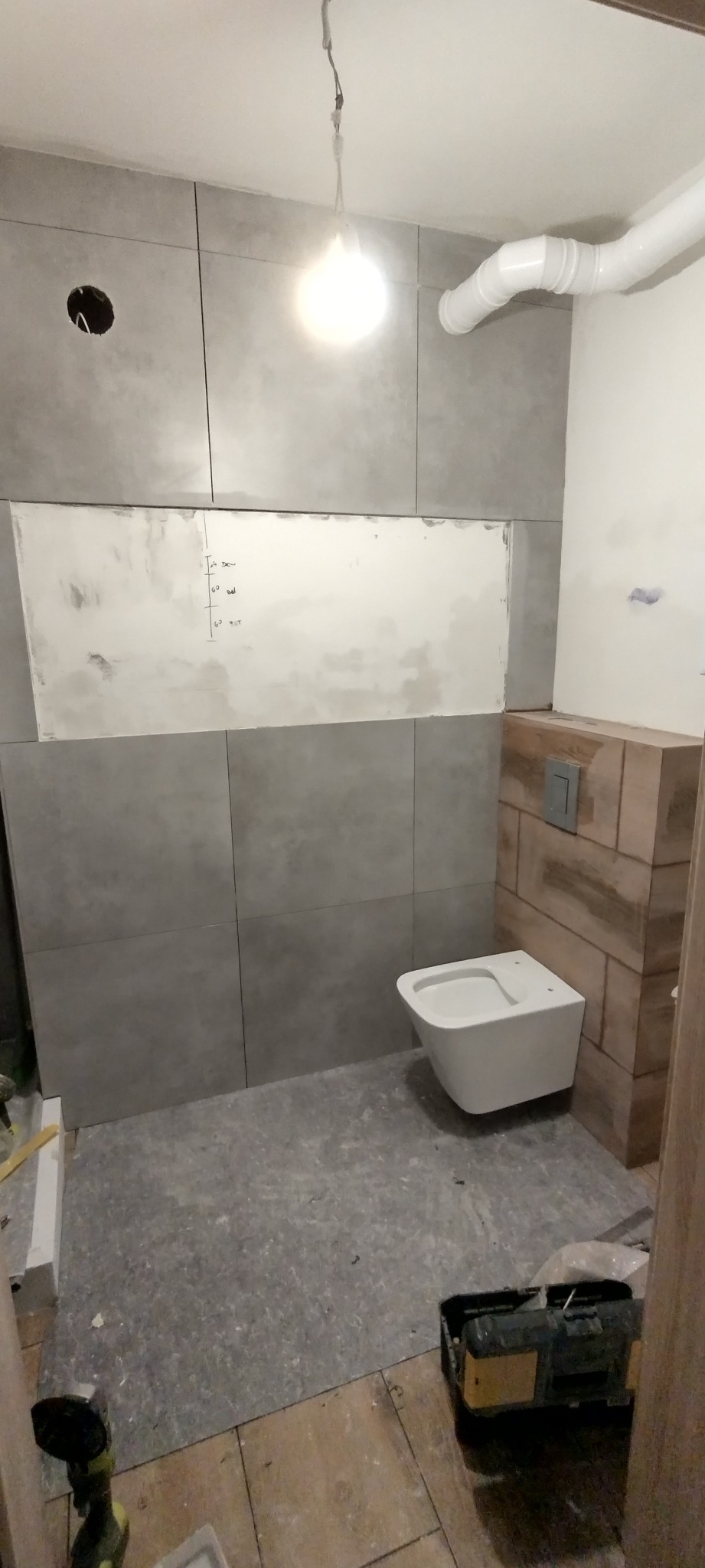 Podwieszana toaleta w trakcie montażu, szare płytki na ścianach, widoczne instalacje i narzędzia, surowy stan wykończenia.