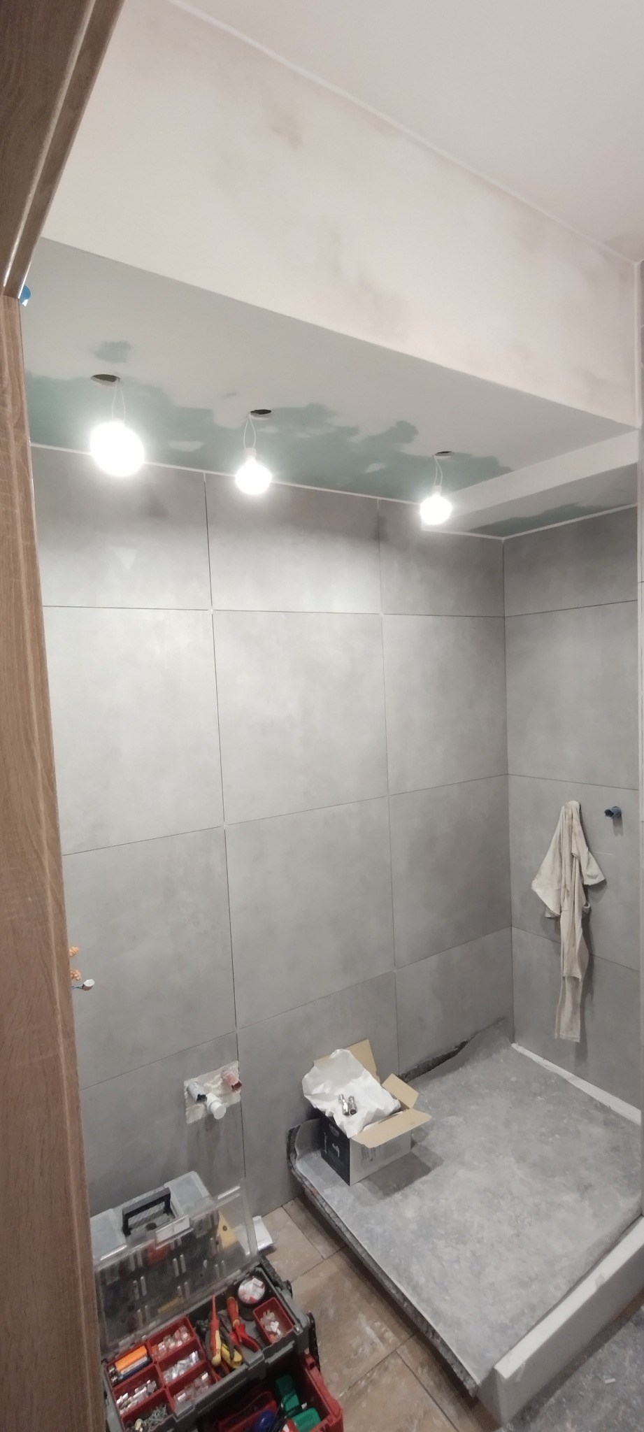 Szare płytki wielkoformatowe na ścianach łazienki w trakcie montażu, widoczne przygotowane podłoże pod prysznic, narzędzia w skrzynce i punktowe oświetlenie sufitowe z niedokończonymi szpachlowaniami.