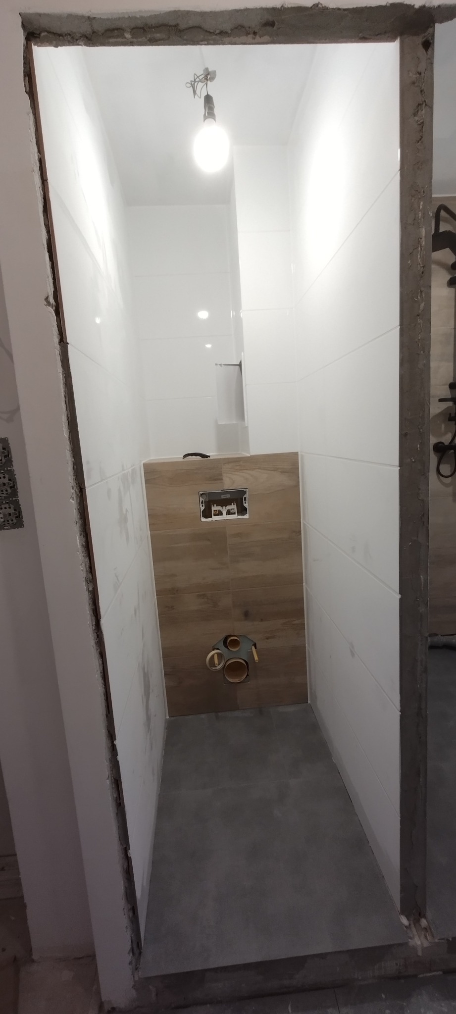 Wnętrze łazienki w trakcie remontu, widoczne białe płytki na ścianach, szare płytki na podłodze oraz drewnopodobna zabudowa stelaża WC z przygotowanymi przyłączami hydraulicznymi, surowa krawędź...