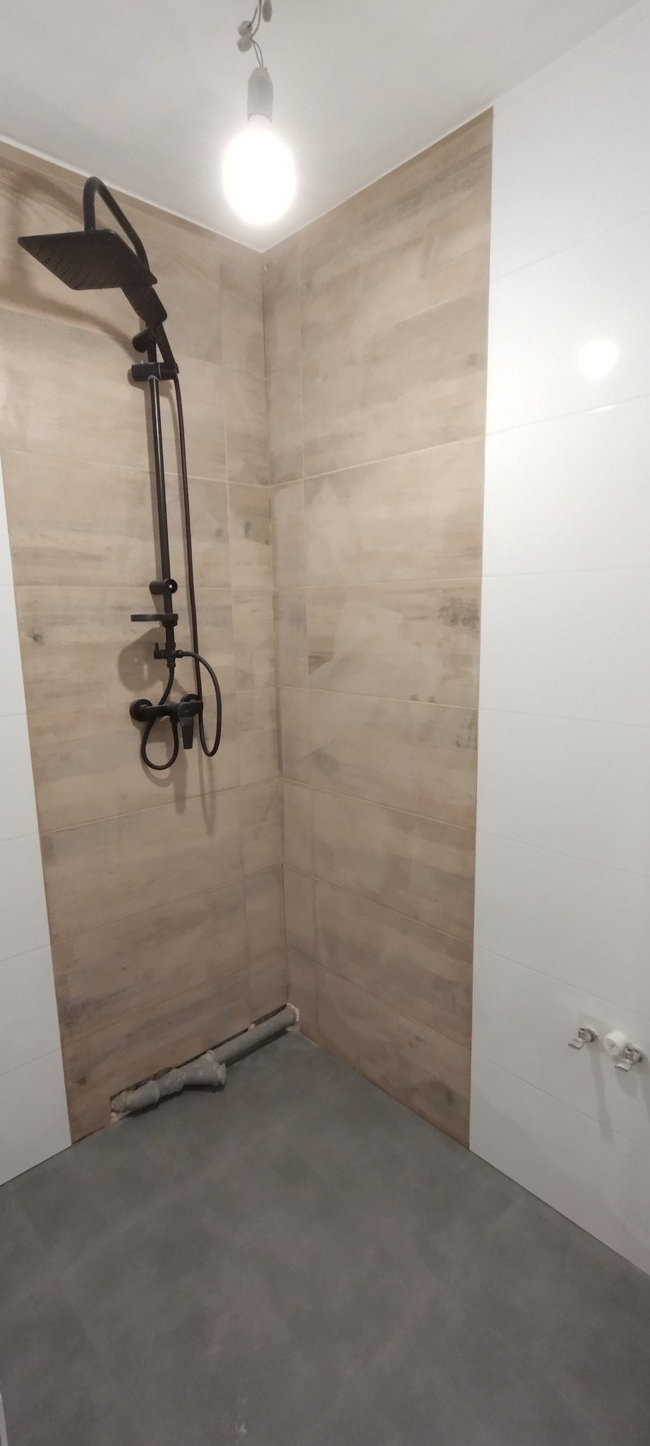 Niedokończona łazienka z zamontowaną czarną armaturą prysznicową, szare płytki podłogowe i częściowo położone płytki ścienne w odcieniach beżu i bieli, widoczne rury odpływowe.