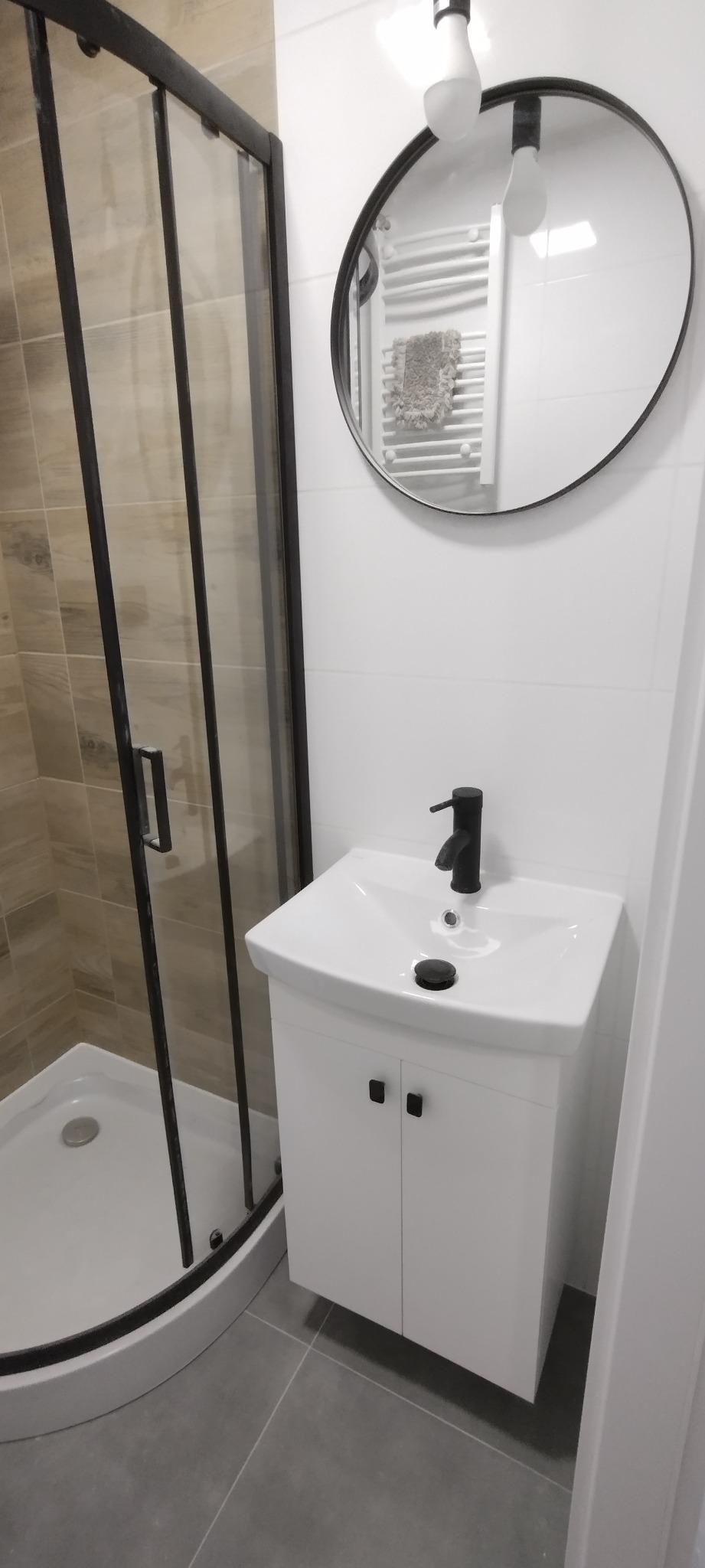Nowoczesna łazienka z kabiną prysznicową z czarnymi ramami, białą szafką pod umywalką z czarną armaturą i okrągłym lustrem z widocznym grzejnikiem drabinkowym w odbiciu.