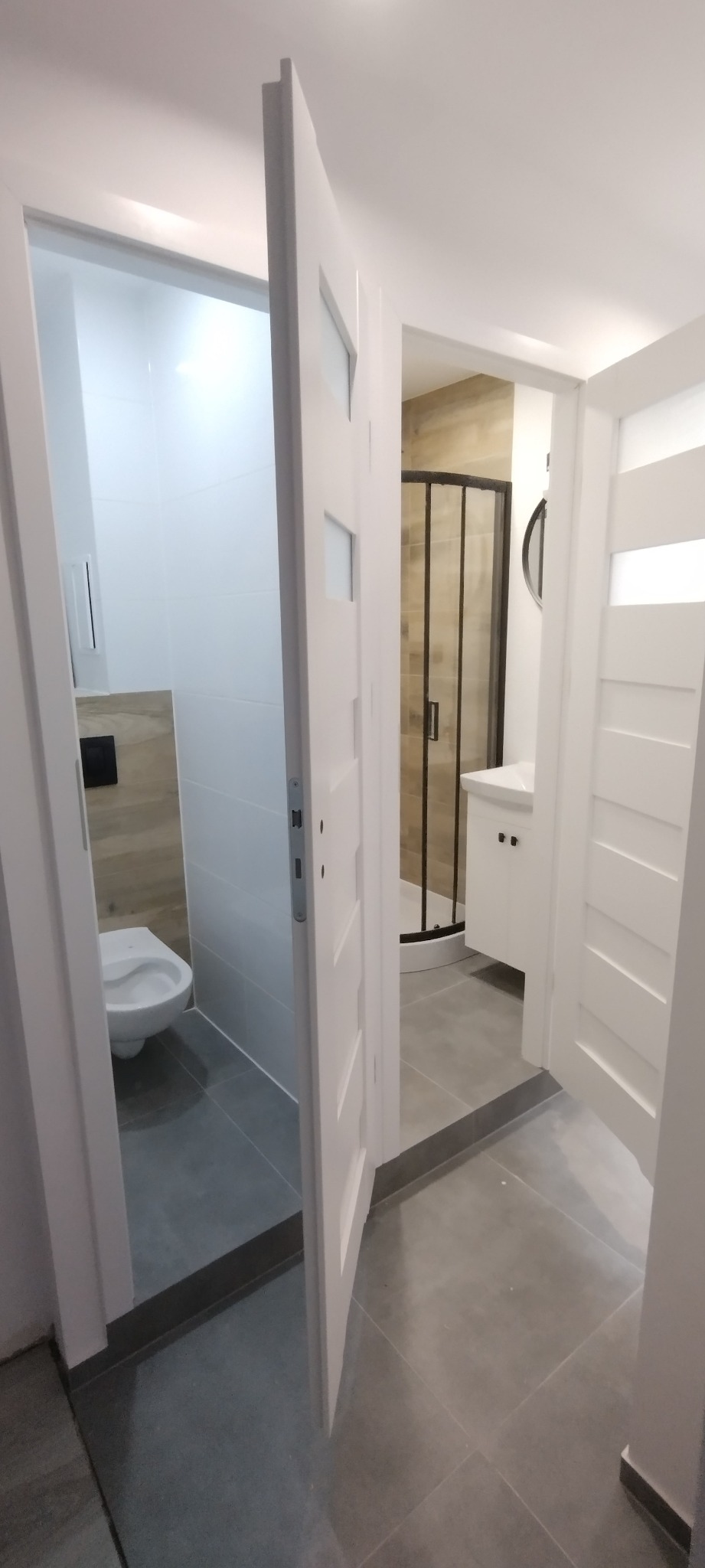 Widok przez otwarte, białe drzwi do dwóch łazienek z szarymi płytkami podłogowymi: jedna z toaletą wiszącą i druga z kabiną prysznicową z czarnymi ramami oraz białą umywalką.