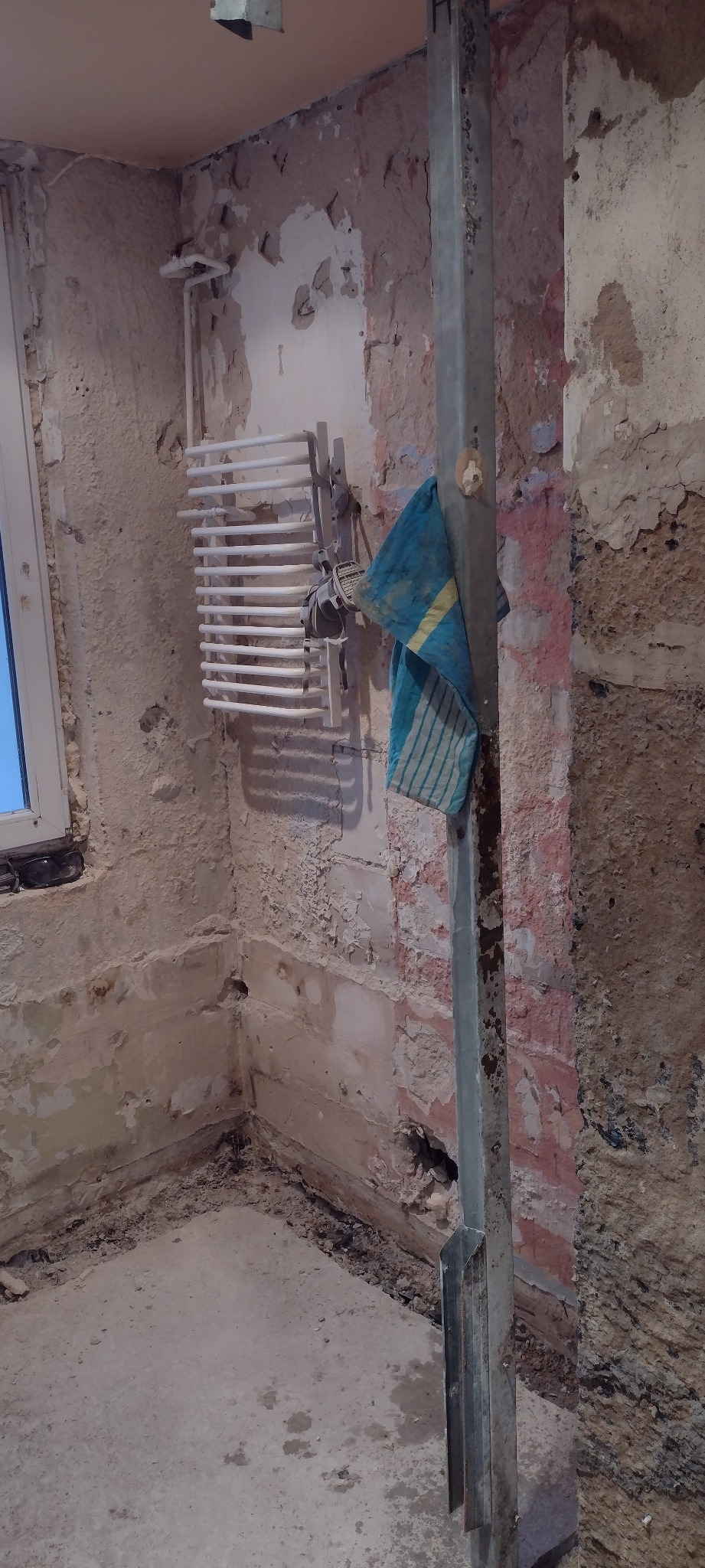 Łazienka w trakcie remontu: ściany z odpadającym tynkiem, grzejnik drabinkowy, pionowa metalowa konstrukcja i niebieska ścierka.