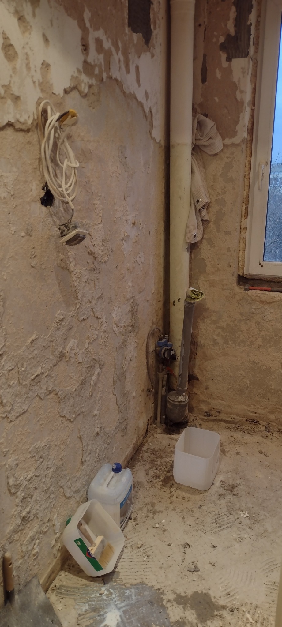 Ściana w trakcie przygotowania do malowania z widocznymi śladami po starych powłokach, odsłonięta instalacja elektryczna, rura pionowa, pojemniki z farbą i narzędzia malarskie na podłodze.