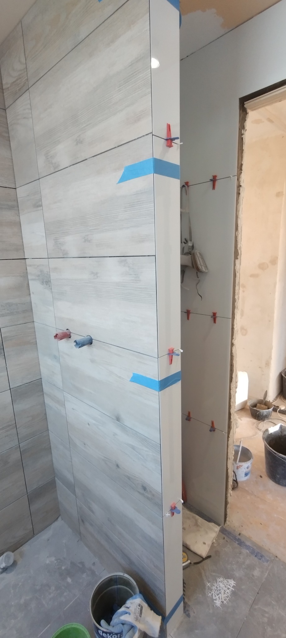 Świeżo ułożone, szare płytki imitujące drewno na ścianie, z użyciem niebieskiej taśmy i klipsów poziomujących, widoczne wiaderka i narzędzia w tle.