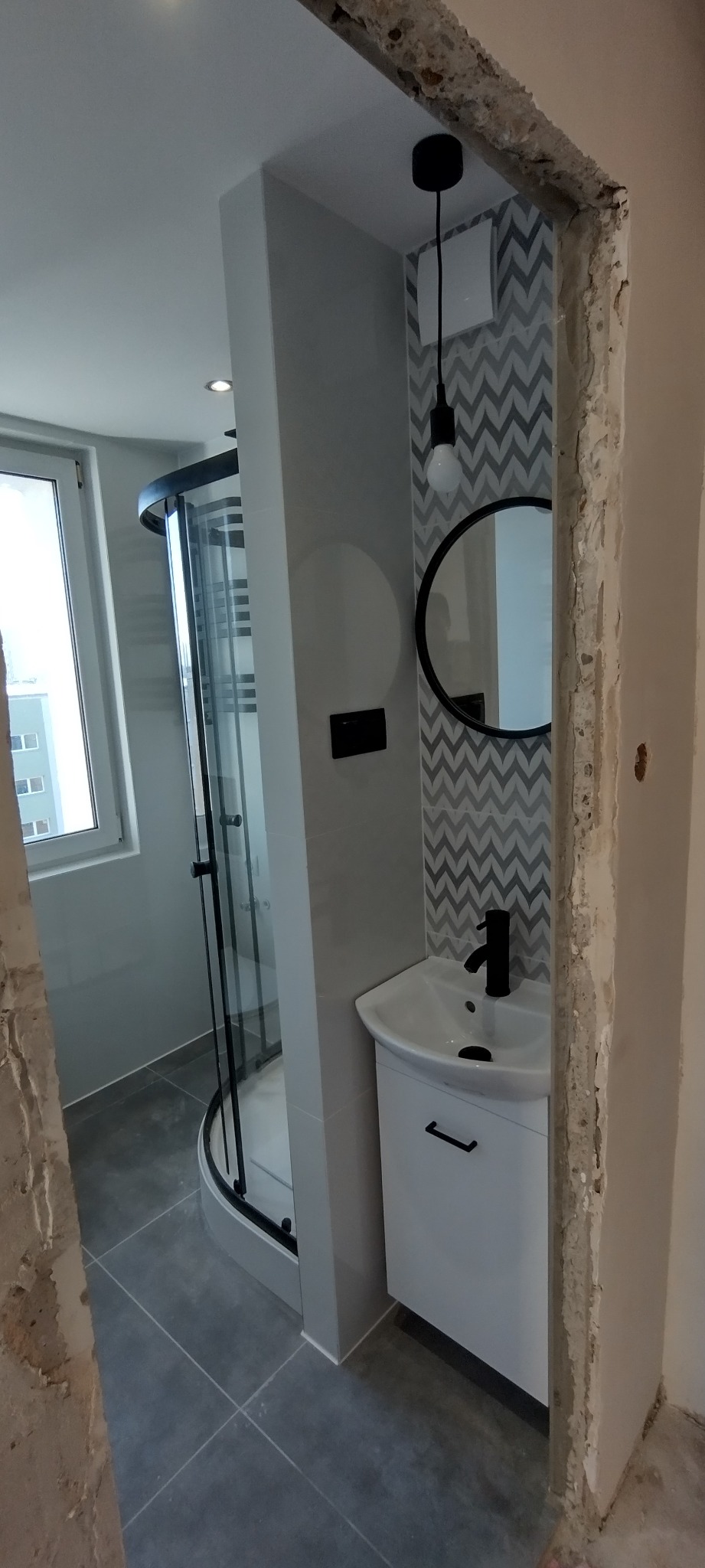 Częściowy widok na wykończoną łazienkę z kabiną prysznicową, umywalką z szafką i okrągłym lustrem, z widocznymi surowymi krawędziami ściany przed wykończeniem.