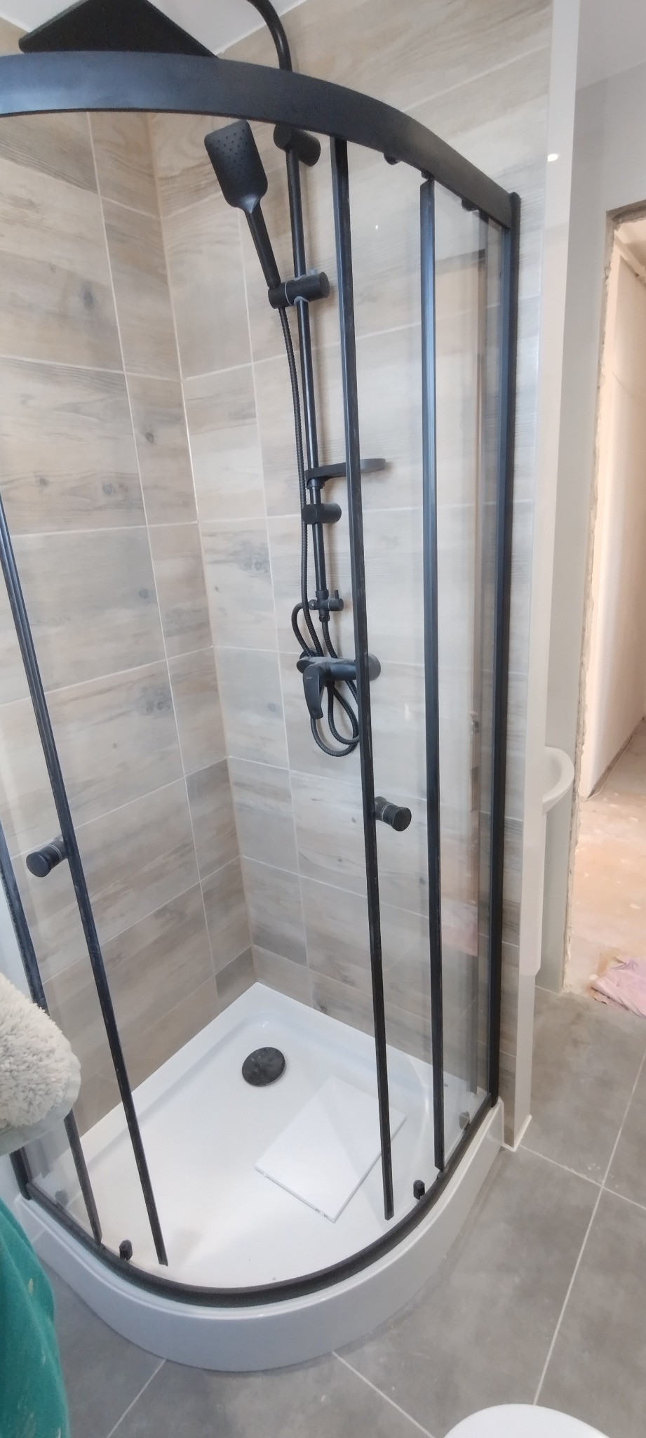 Kabina prysznicowa z czarnymi profilami i deszczownicą zamontowana w wykończonej łazience z jasnymi, drewnopodobnymi płytkami na ścianach.