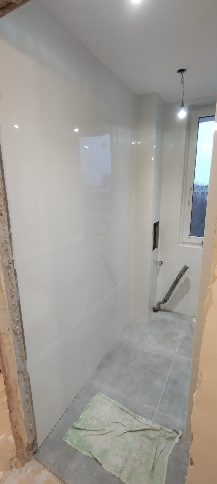 Wnętrze łazienki w trakcie prac wykończeniowych: białe płytki ścienne z połyskiem, szare płytki podłogowe, widoczny fragment instalacji wodnej, okno i fragment surowej ściany z resztkami zaprawy...